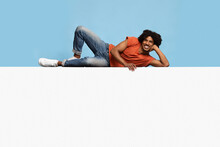 Happy Black Guy Relaxing On Huge Empty Placard Over Blue