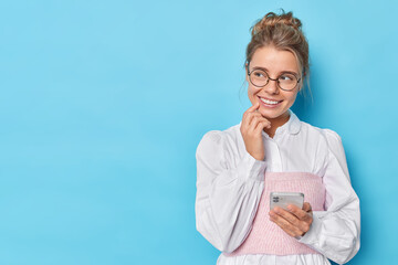 Wall Mural - Pensive smiling young woman holds mobile phone and thinks about something pleasant looks away wears optical glasses and white elegant shirt isolated over blue studio background. Technology user