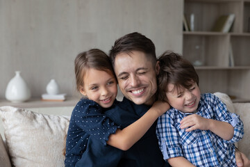 Wall Mural - Son and daughter cuddling young cheerful daddy. Loving family sit on sofa embracing laughing feel excited, dad enjoy caress and love of kids, receive congratulations, hugs on Happy Father Day concept