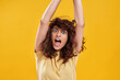 Portrait of amazed young woman with dark curly hair in casual wear looking surprised at camera with arms raised isolated over yellow background