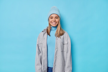 Wall Mural - Horizontal shot of good looking woman in hat and grey jacket smiles positively has happy mood isolated over blue background ready for walk. Cheerful millennial girl in outerwear poses indoor