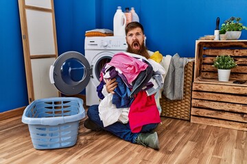 Sticker - Redhead man with long beard putting dirty laundry into washing machine in shock face, looking skeptical and sarcastic, surprised with open mouth