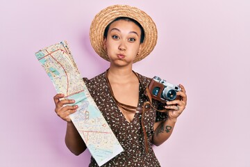 Wall Mural - Beautiful hispanic woman with short hair holding city map and vintage camera puffing cheeks with funny face. mouth inflated with air, catching air.