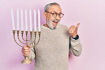 Poster - Handsome senior man with beard holding menorah hanukkah jewish candle pointing thumb up to the side smiling happy with open mouth