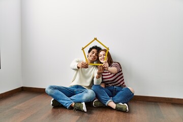 Wall Mural - Young hispanic couple holding house project sitting on the floor at empty new home.