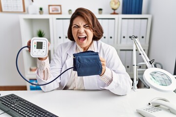 Poster - Middle age hispanic doctor woman using blood pressure monitor angry and mad screaming frustrated and furious, shouting with anger. rage and aggressive concept.