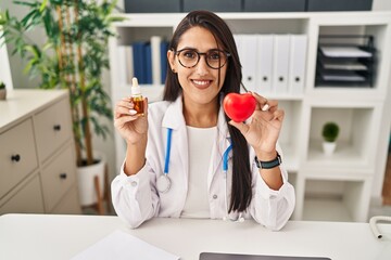 Wall Mural - Young hispanic doctor woman holding heart and cbd oil smiling with a happy and cool smile on face. showing teeth.