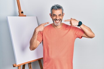 Poster - Handsome middle age man with grey hair standing by painter easel stand smiling cheerful showing and pointing with fingers teeth and mouth. dental health concept.