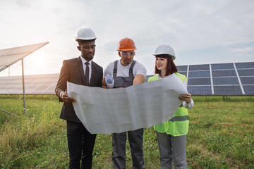 Wall Mural - Two engineers and one technician examining drawings of solar farm while standing together outdoors. Male and female partners in helmets working on modern business project.