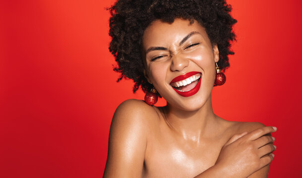 Portrait of beautiful black woman with afro curly hairstyle, red lipstick and glowing healthy skin, New Year eve toy earrings, laughing and smiling. Concept of winter promo sale