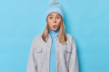 Wall Mural - Portrait of shocked young European woman impressed by big announcement stares in awewears knitted hat and grey jacket isolated over blue background. Overwhelmed female model feels concerned.