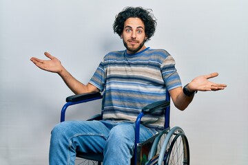 Wall Mural - Handsome hispanic man sitting on wheelchair smiling showing both hands open palms, presenting and advertising comparison and balance