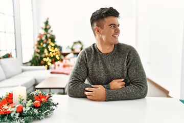 Sticker - Young hispanic man sitting on the table by christmas tree looking away to side with smile on face, natural expression. laughing confident.