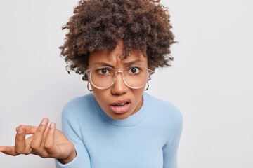 Canvas Print - Puzzled indignant woman says so what raises hand and looks displeased at camera frowns face with dissatisfactionn wears big transparent glasses casual blue jumper isolated over white background