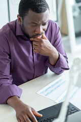 Financial report. Puzzled male worker. Difficult task. Concentrated man thinking about paper documents working laptop in light office interior.