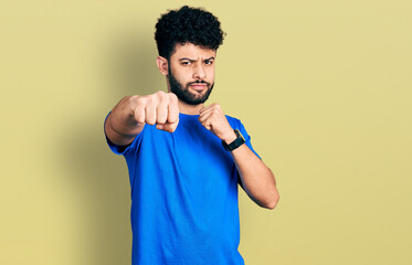 Sticker - Young arab man with beard wearing casual blue t shirt punching fist to fight, aggressive and angry attack, threat and violence