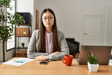 Sticker - Young chinese business worker wearing business style sitting on desk at office with serious expression on face. simple and natural looking at the camera.