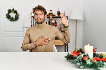 Poster - Young handsome man with beard sitting on the table by christmas decoration swearing with hand on chest and open palm, making a loyalty promise oath