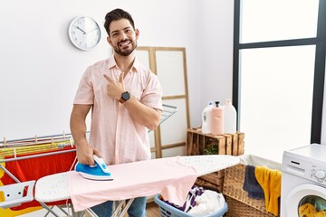 Poster - Young man with beard ironing clothes at home cheerful with a smile on face pointing with hand and finger up to the side with happy and natural expression