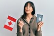 © Krakenimages.com - Young hispanic woman holding canada flag and passport smiling looking to the side and staring away thinking.