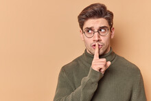 Be Quiet. Serious Mysterious Man Presses Index Finger To Lips Shows Taboo Sign Tells To Keep Silence Shushing Indoor Wears Casual Clothes Looks Away Isolated Over Beige Background Blank Copy Space