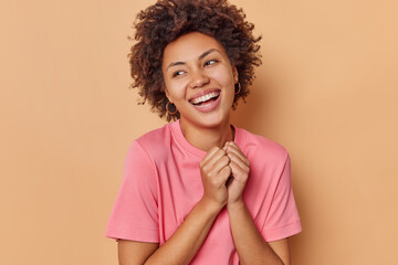 Poster - Photo of carefree pretty woman keeps hands together smiles broadly and looks away expresses positive emotions wears casual pink t shirt isolated over beige background admires something nice.