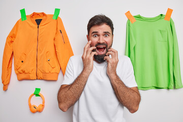 Wall Mural - Photo of panicking adult man stares with scared expression exclaims emotionally keeps hands on face wears casual t shirt poses indoor against items of clothes. Bearded guy reacts on something