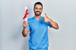 Handsome hispanic man with beard wearing blue male nurse uniform holding diploma smiling happy and positive, thumb up doing excellent and approval sign