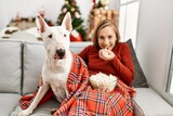 Fototapeta Psy - Young caucasian woman watching movie sitting with dog by christmas tree at home