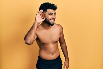 Canvas Print - Arab man with beard wearing swimwear shirtless smiling with hand over ear listening an hearing to rumor or gossip. deafness concept.