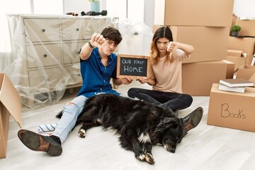 Canvas Print - Young caucasian couple with dog holding our first home blackboard at new house looking unhappy and angry showing rejection and negative with thumbs down gesture. bad expression.