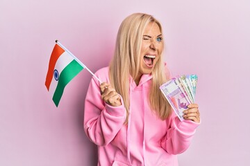 Poster - Young blonde woman holding india flag and rupee banknotes angry and mad screaming frustrated and furious, shouting with anger. rage and aggressive concept.