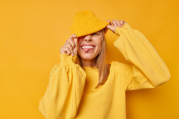 Wall Mural - Joyful happy woman hides eyes with hat sticks out tongue has playful expression teases someone wears casual jumper isolated over yellow background squints happily shows true emotions. People and fun