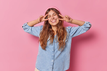 Wall Mural - Young happy woman sends positive vibes makes peace gesture over eyes smiles toothily dressed in denim shirt isolated over pink background shows victory sign has cheerful mood. Body language.