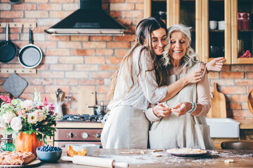 Young woman and mother in white aprons stand at large brown wooden kitchen table and prepare tasty dinner. Senior mother and happy adult daughter baking pumpkin pie together at home.