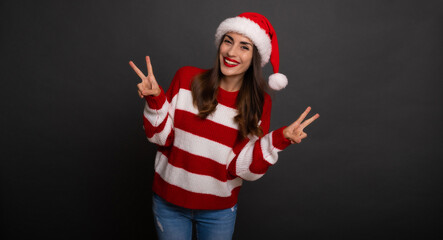 Wall Mural - Excited beautiful smiling happy young woman in Christmas Santa hat shows the Victory sign and looks on camera