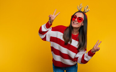 Wall Mural - Close up portrait of an attractive excited smiling young girl in Christmas reindeer antlers and sweater while she posing isolated on yellow background in studio