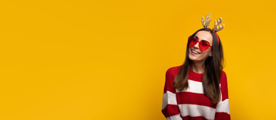 Wall Mural - Banner wide photo of an attractive excited smiling young girl in Christmas reindeer antlers and sweater while she posing isolated on yellow background in studio