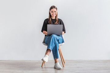 Poster - Happy young woman working online, sitting on chair and using laptop against white studio wall, full length