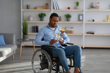 Wall Mural - Unhappy black paraplegic sportsman in wheelchair holding trophies, feeling depressed indoors