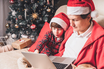 Father with child kid son and puppy dog in Santa hat at Christmas holidays, sitting on a couch, having a video chat on laptop in the living room with Christmas tree at home. Boys watch movie