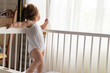 © Ivanka - Cute little 1 years happy toddler baby girl kid sleeping sweetly in white crib in white pajama with pillow infant child look out big window on hot summer day at home. Childhood concept. copy space