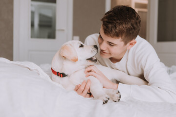 portrait of a teenage boy lying in bed on white bedding in an embrace with a light-colored dog. boy is fooling around with a pet in the room. space for text. High quality photo