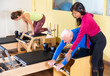 Elderly man practicing Pilates system exercises on wunda chair under guidance of positive qualified Hispanic female trainer