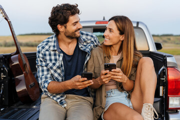 Wall Mural - White couple smiling and using cellphones while sitting in car trunk