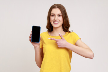 Wall Mural - Look at my cellphone! Adorable glad teenager girl pointing smartphone with finger and smiling at camera, advertising of mobile device. Indoor studio shot isolated on gray background.