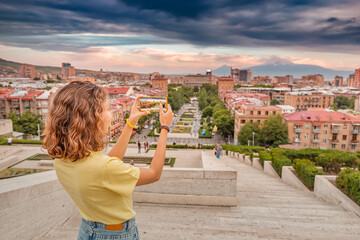 Happy woman taking pictures of a scenic urban landscape in Yerevan city with a distant view of famous Mount Ararat and Cascade complex