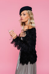 Wall Mural - stylish woman in black beret and faux-fur jacket looking at camera while holding cocktail isolated on pink