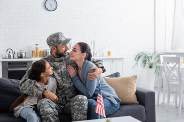 Poster - Man in camouflage uniform hugging family near american flag at home