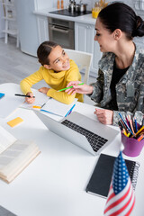 Poster - Smiling mother in military uniform holding pencil near daughter with notebook and laptop at home